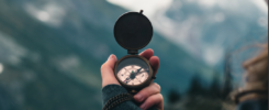 A person holding an open compass, facing cloudy mountain peaks